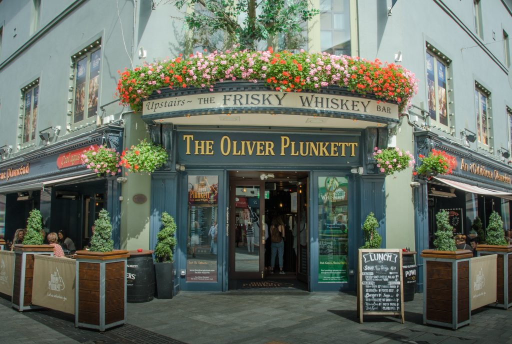 Corner frontage of The Oliver Plunkett, a traditional Irish pub on Oliver Plunkett Street in Cork city: a navy-blue shopfront with gold lettering, a flower box of red and pink geraniums over the entrance, an 'Upstairs - The Frisky Whiskey B