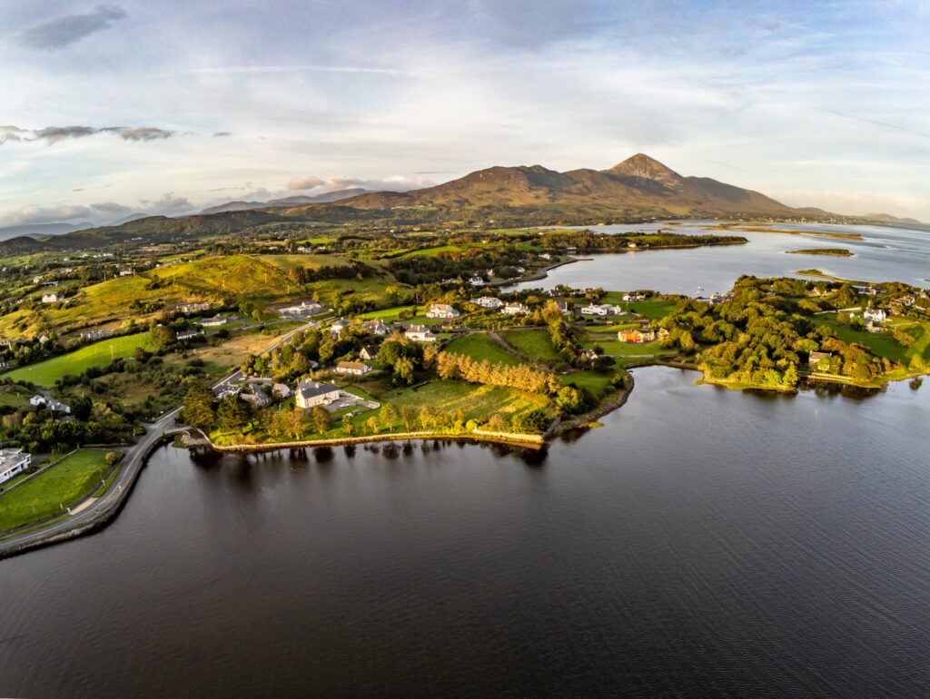 Aerial drone view over Westport Bay (Clew Bay) near Westport, County Mayo, Ireland in warm low sunlight: calm dark water wraps around a green peninsula of fields, hedgerows, houses and autumn-tinged trees with a coastal road, while the cone